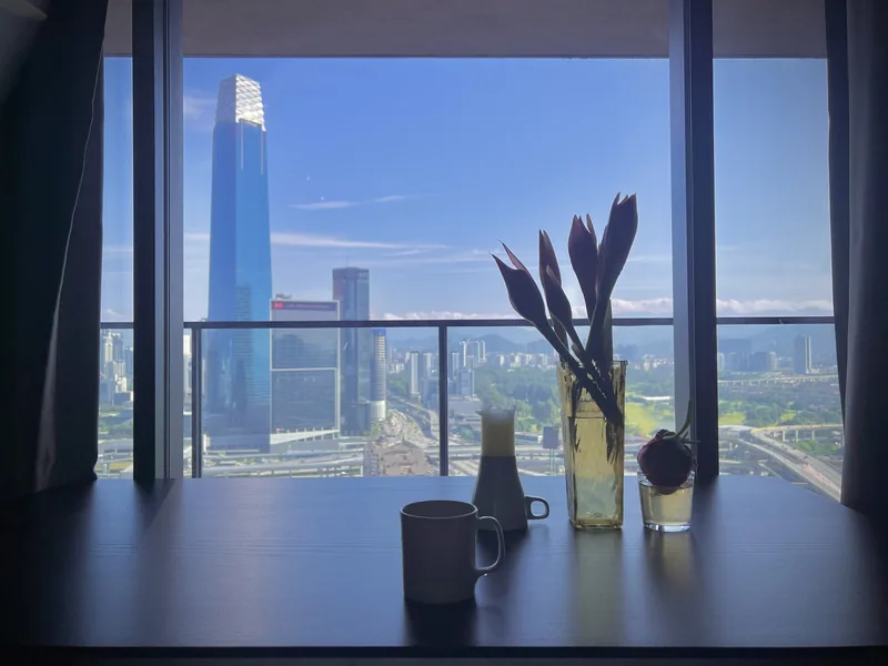 Bàn khách sạn với ly và bình hoa nhìn ra đường chân trời Kuala Lumpur qua khung kính lớn.