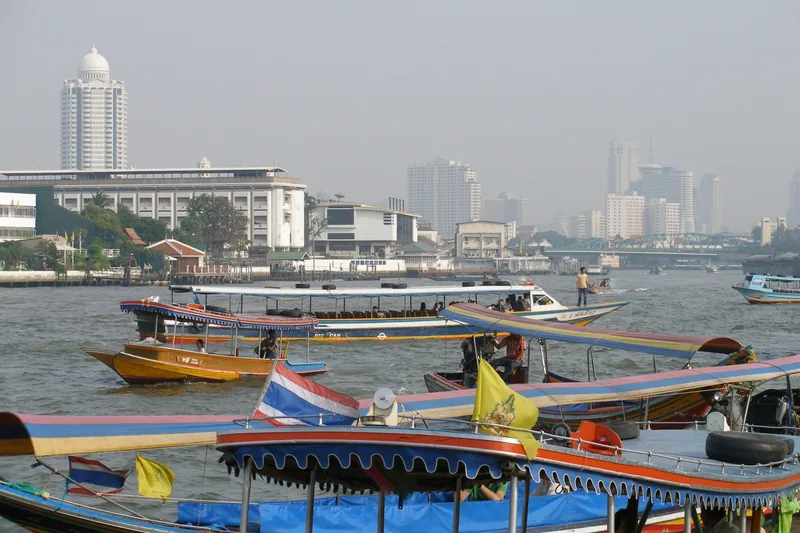 Những thuyền đuôi dài cắm cờ Thái Lan tấp nập trên sông Chao Phraya với đường chân trời Bangkok mờ sương phía sau.
