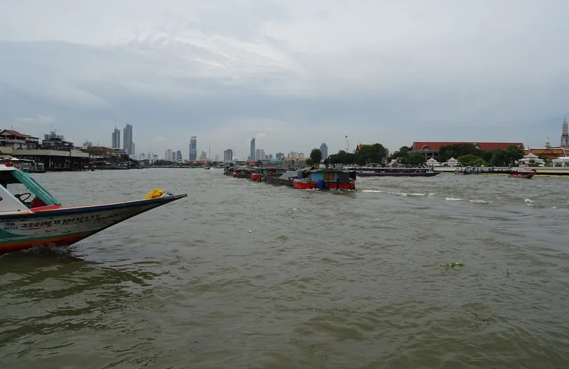 Một chiếc thuyền đuôi dài và các sà lan lướt trên sông Chao Phraya rộng lớn với đường chân trời Bangkok phía xa.