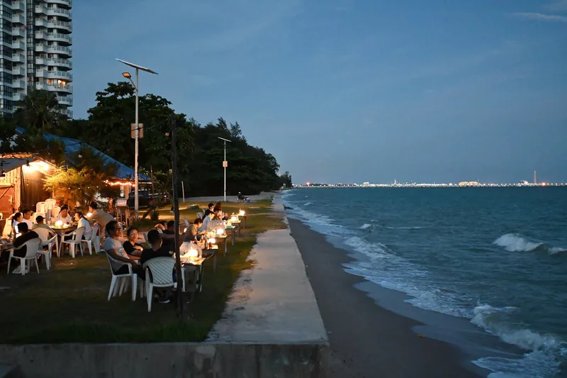 Thực khách dùng bữa bên những bàn nến dọc bờ biển lúc chạng vạng, sóng vỗ nhẹ vào bờ.
