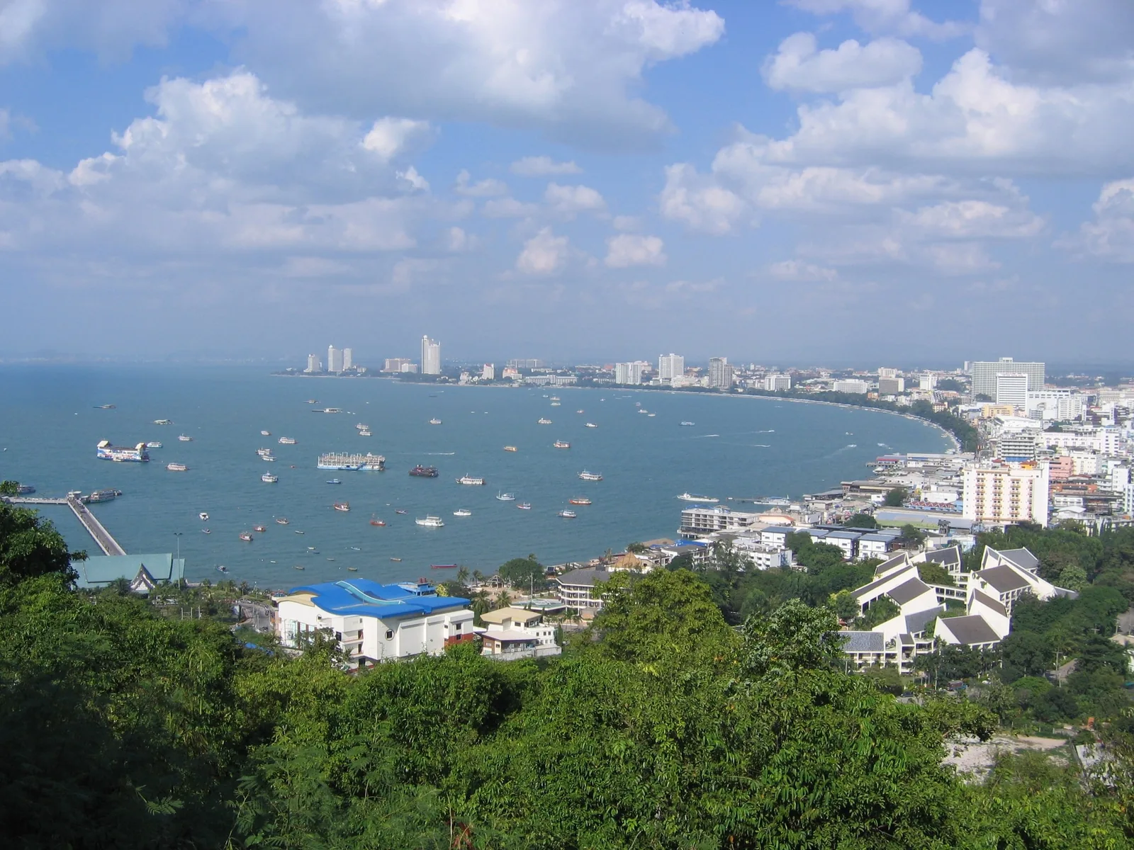 Toàn cảnh vịnh Pattaya với bãi biển cong, thuyền bè trên biển và các tòa nhà thành phố nhìn từ đỉnh đồi cây xanh.
